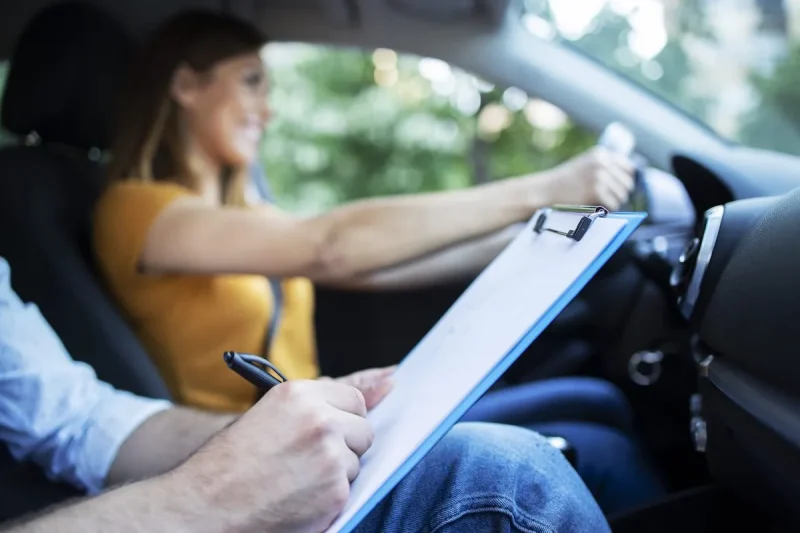 close-up-view-driving-instructor-holding-checklist-while-background-female-student-steering-driving-car-scaled
